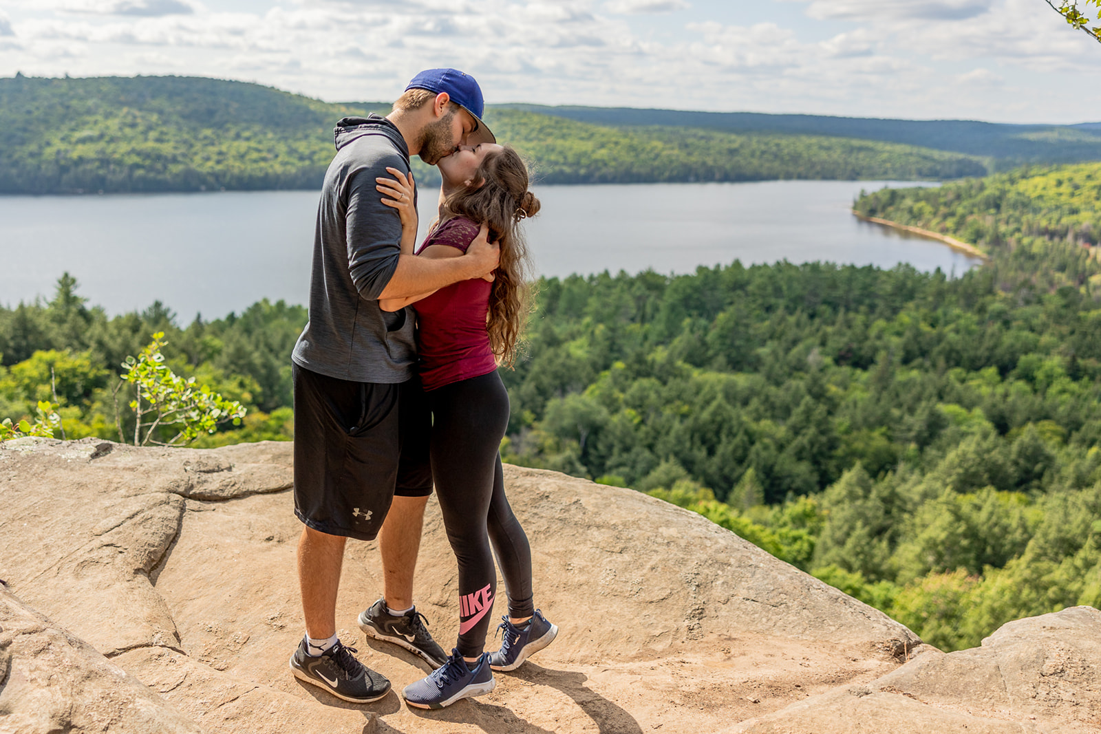 Newly engaged couple kissing at the top of Booth's Rock trail in Algonquin Provincial Park with Rock Lake and the surrounding forest stretching behind them, photographed by Kitchener-Waterloo proposal photographer Alexia Cina.