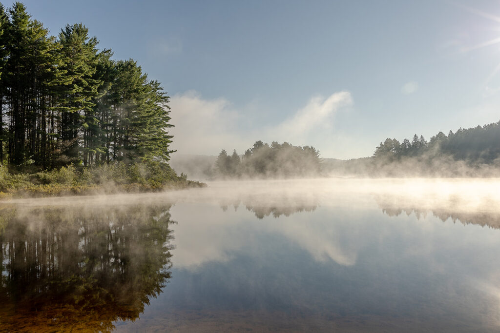 Morning mist rising off a lake in Algonquin Provincial Park at sunrise, trees reflected in the still water.